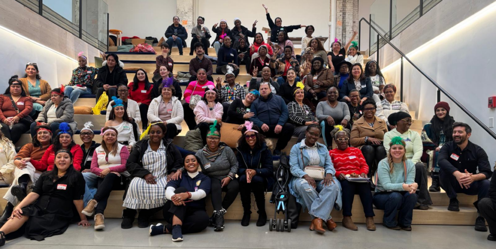 A large, diverse group of people sit and stand on tiered wooden seating indoors, facing the camera and smiling.