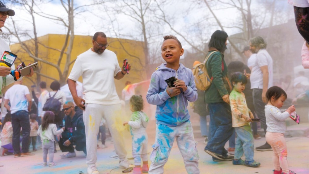Children and adults participate in an outdoor color powder festival, wearing casual clothes stained with colorful powder, surrounded by trees and other people.