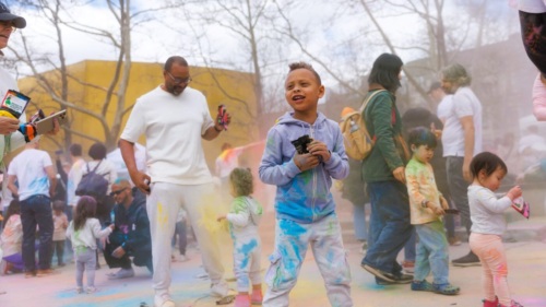 Children and adults participate in an outdoor color powder festival, wearing casual clothes stained with colorful powder, surrounded by trees and other people.
