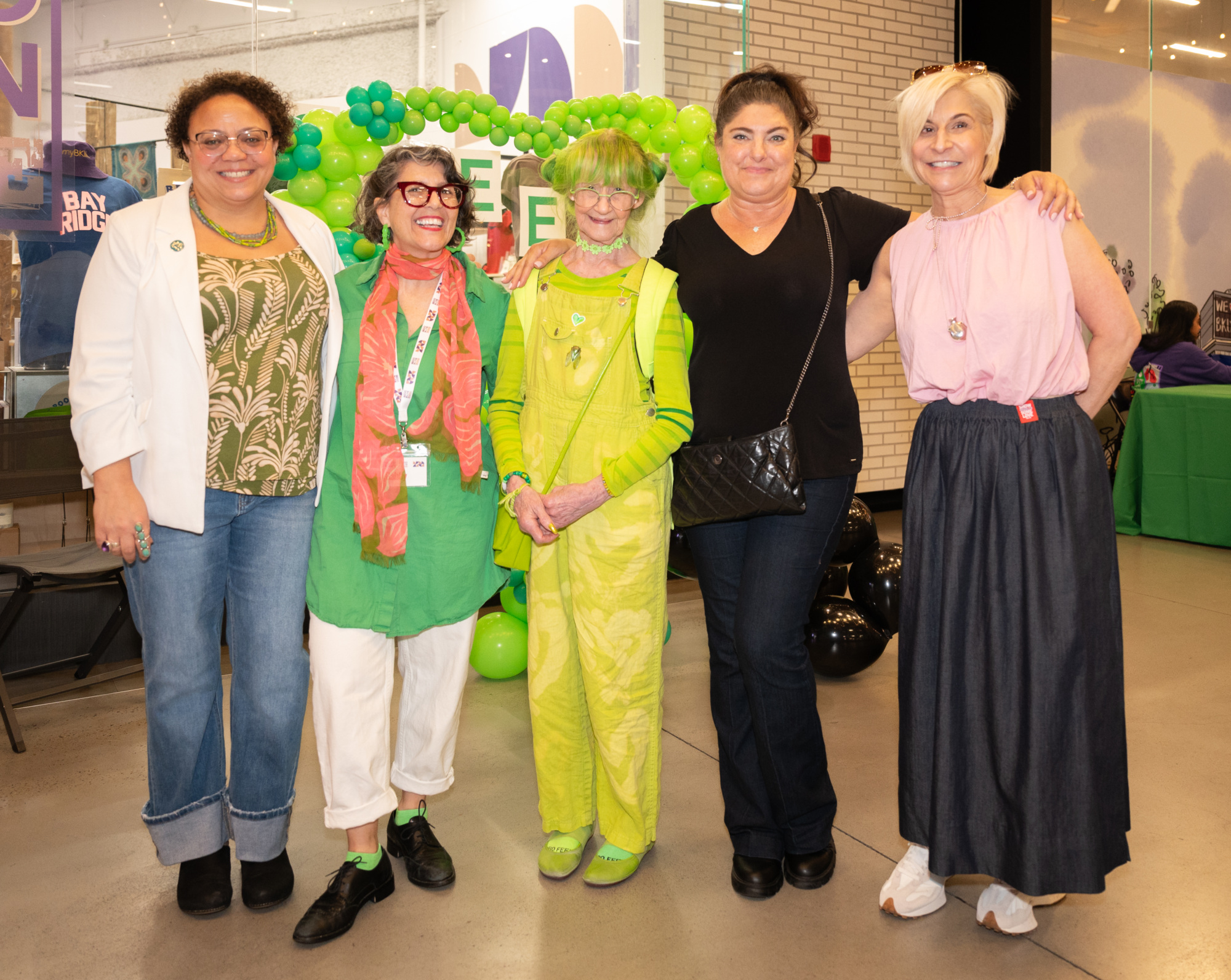 Five women stand together indoors, smiling for a group photo; two are dressed in bright green outfits while the others wear casual attire. Green balloons and event decor are visible in the background.