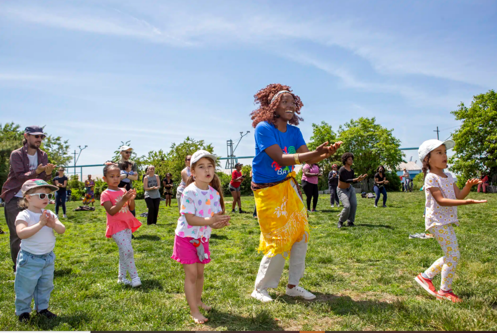 A group of children and adults stand on grass, clapping and following along to instructions at an outdoor event on a sunny day.