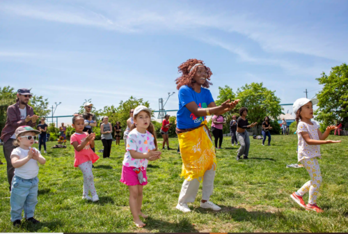A group of children and adults stand on grass, clapping and following along to instructions at an outdoor event on a sunny day.