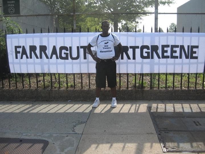 A man stands on a sidewalk in front of a large banner reading "FARRAGUT FORT GREENE" and wears a shirt that says "Old Timers Reunion.