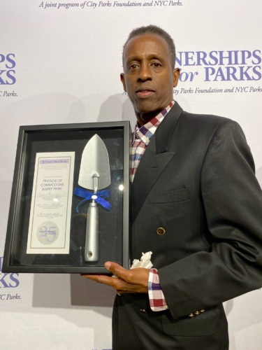 A man stands in front of a "Partnerships for Parks" backdrop holding a framed certificate and ceremonial silver trowel award.