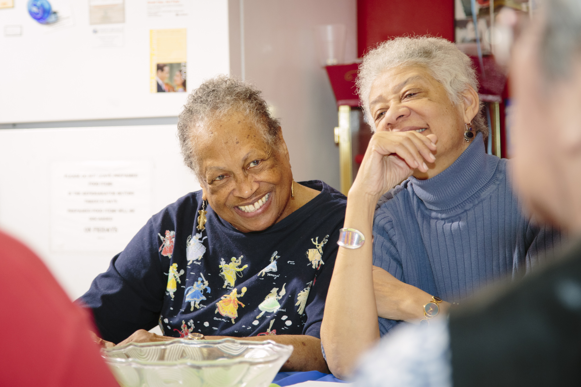 Two elderly women sit at a table, smiling and engaged in conversation with others in a brightly lit room.