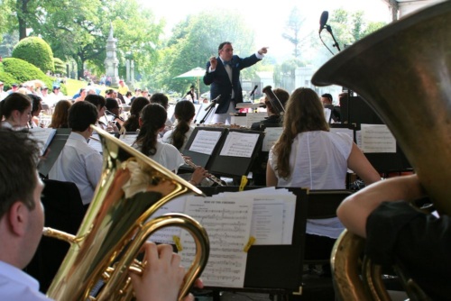 A conductor leads an outdoor orchestra performance, with musicians playing brass and woodwind instruments and sheet music visible on stands.