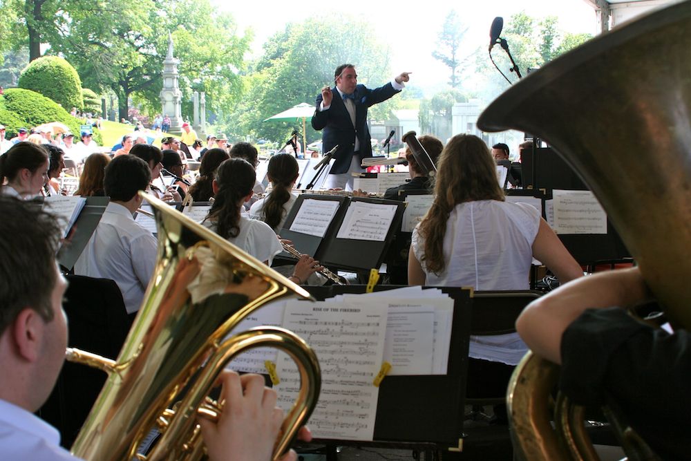 A conductor leads an outdoor orchestra performance, with musicians playing brass and woodwind instruments and sheet music visible on stands.