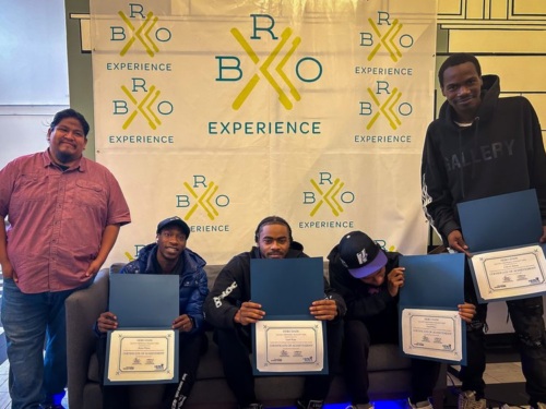 Five people pose in front of a "BKO Experience" banner; four are seated holding certificates, while one stands to the left without a certificate, all facing the camera.