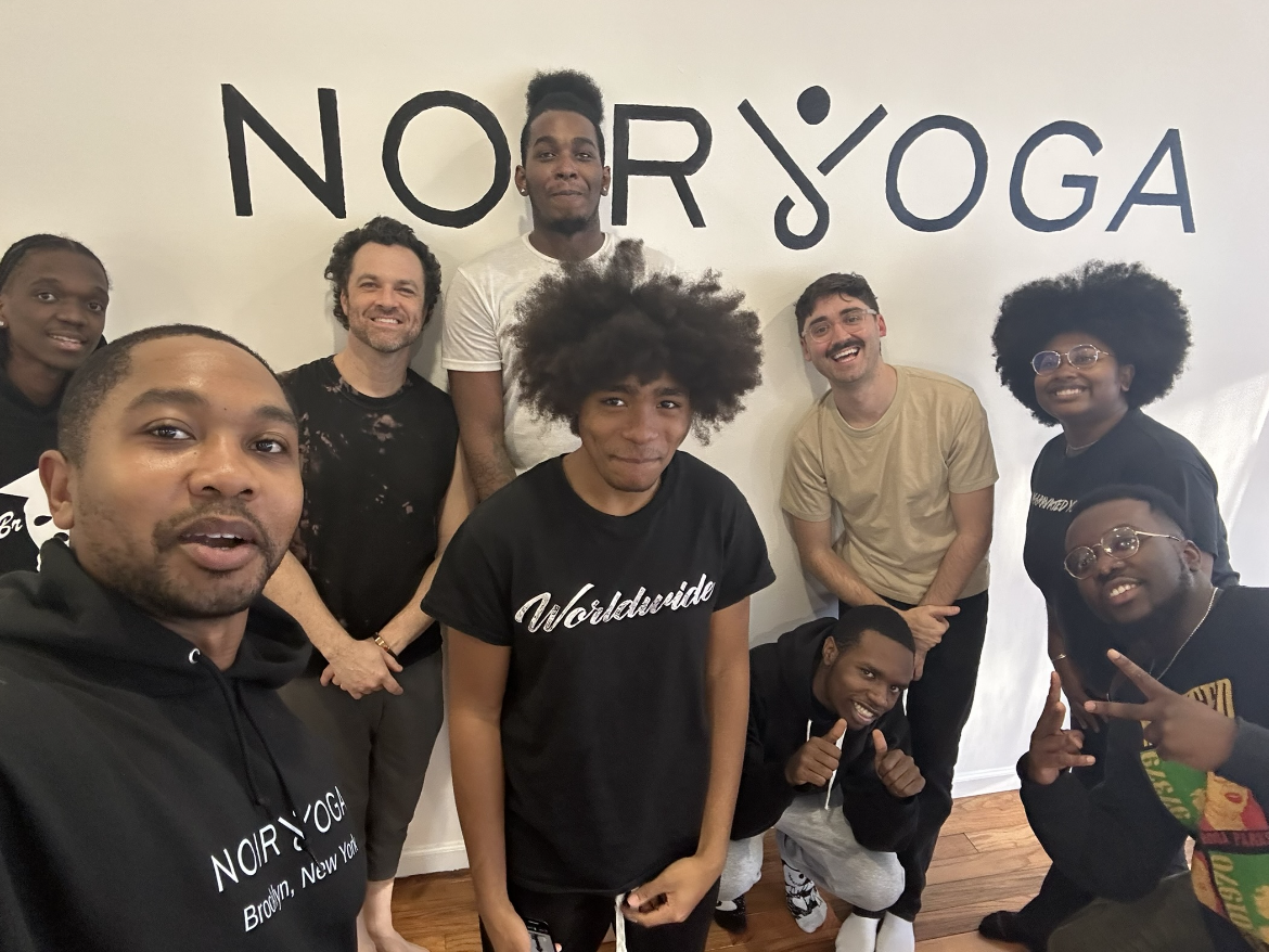 Nine people pose and smile in front of a wall with the text "NORRYOGA." Some are standing, others crouching, in casual attire, indoors on a wood floor.