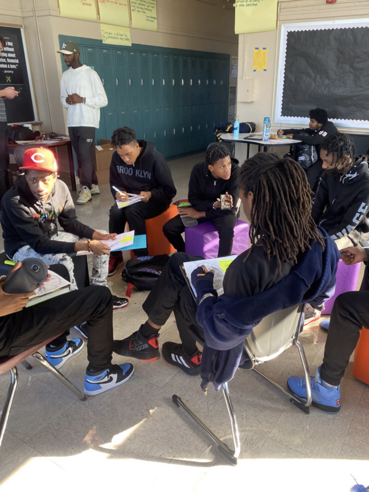 A group of students sits in a classroom circle, reading papers and discussing, with another person standing in the background near lockers.