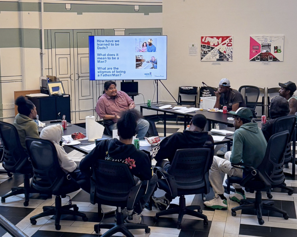 A group of people sit in a circle around tables, listening to a speaker. A screen displays discussion questions about fatherhood. Posters and diagrams are visible on the walls.