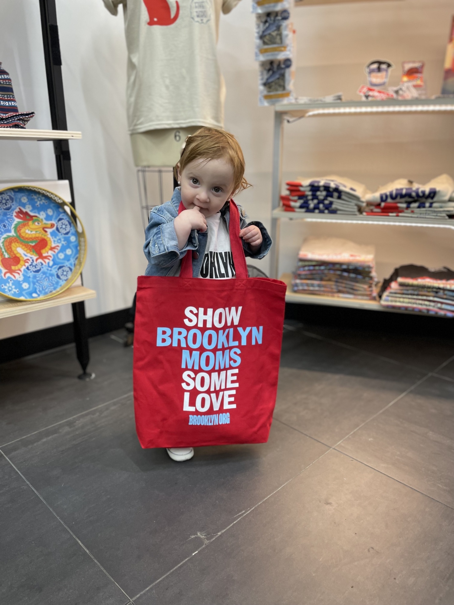 A small child stands indoors holding a large red tote bag that reads “SHOW BROOKLYN MOMS SOME LOVE,” with folded shirts on shelves in the background.