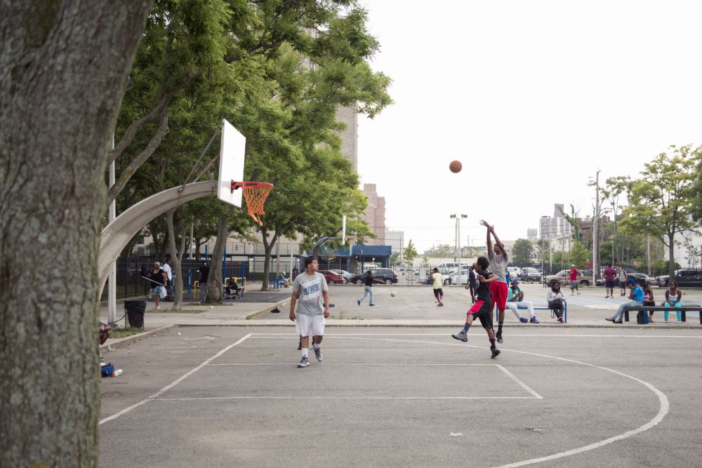 People play basketball on an outdoor court; one player jumps to take a shot while others watch or play nearby, with trees and buildings in the background.
