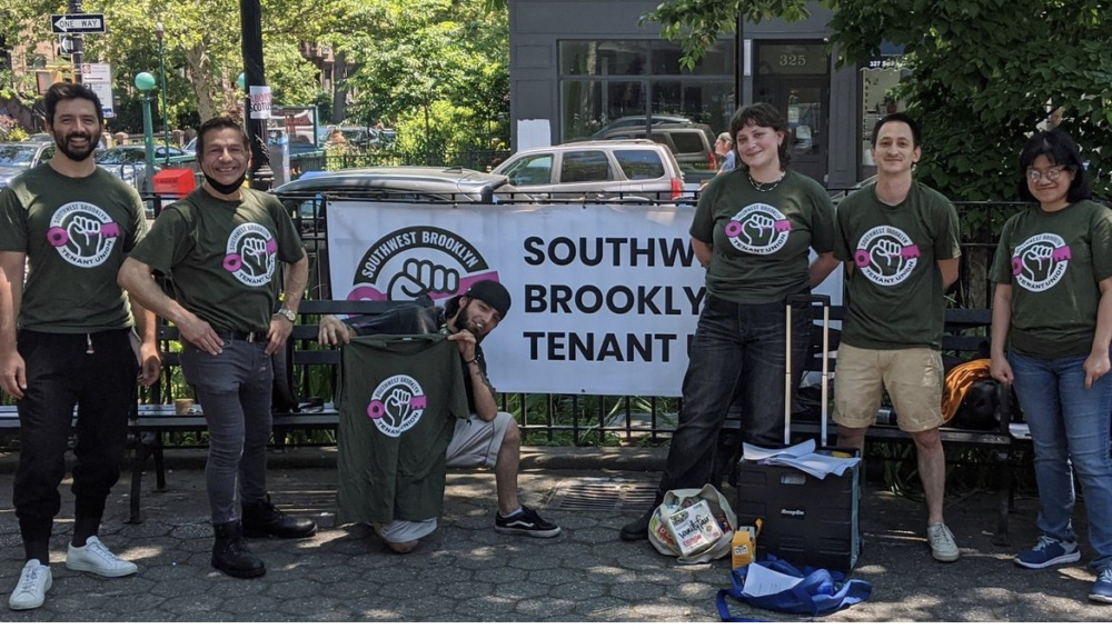 Six people wearing matching green shirts with a fist logo stand in front of a "Southwest Brooklyn Tenant" banner at an outdoor event with tables and supplies.
