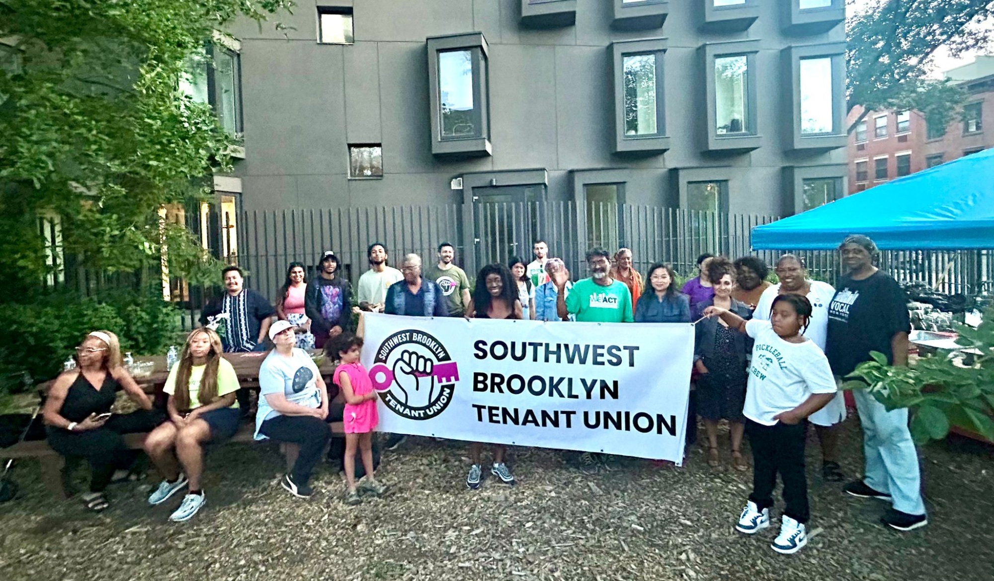 A group of people stands outdoors holding a banner that reads "Southwest Brooklyn Tenant Union" in front of a building, with trees and a blue canopy nearby.