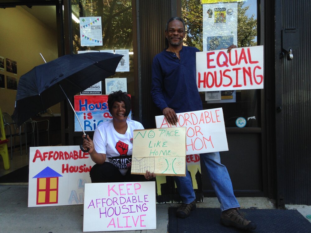 Two people stand outside a building holding signs advocating for affordable and equal housing, including messages like "KEEP AFFORDABLE HOUSING ALIVE" and "EQUAL HOUSING.