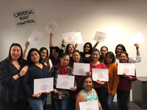 A group of women stand and sit together indoors, holding certificates and smiling; a sign on the wall reads “Universal Rent Control.”.