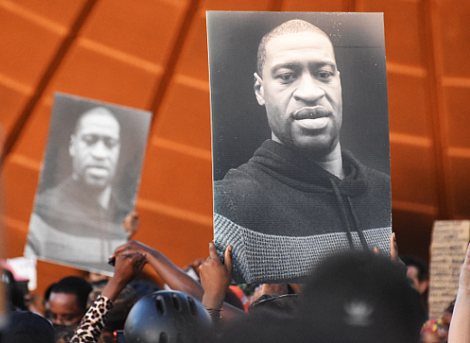 People hold up large black-and-white portraits of George Floyd at a public gathering or protest.