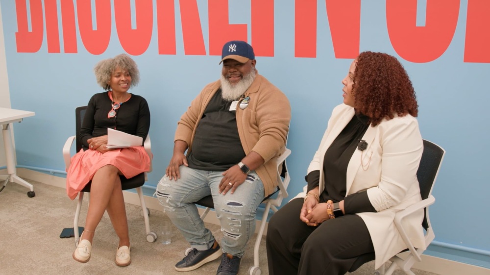 Three people sit and talk in an office setting with a blue wall and large red letters in the background. They appear to be engaged in a friendly conversation.