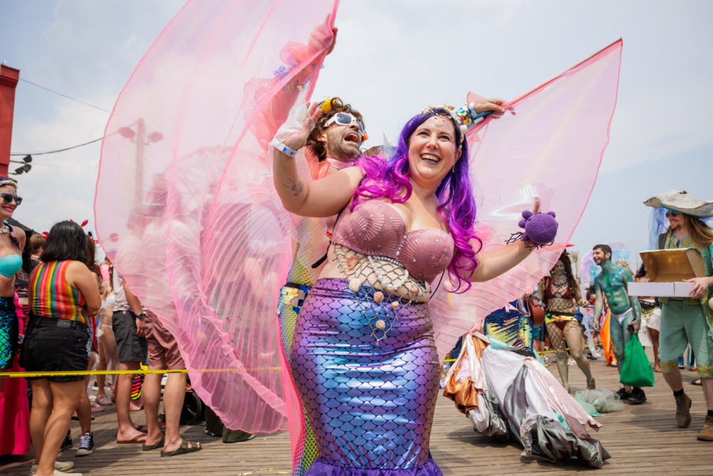 A group of people in colorful mermaid costumes participate in a festive outdoor event on a boardwalk.