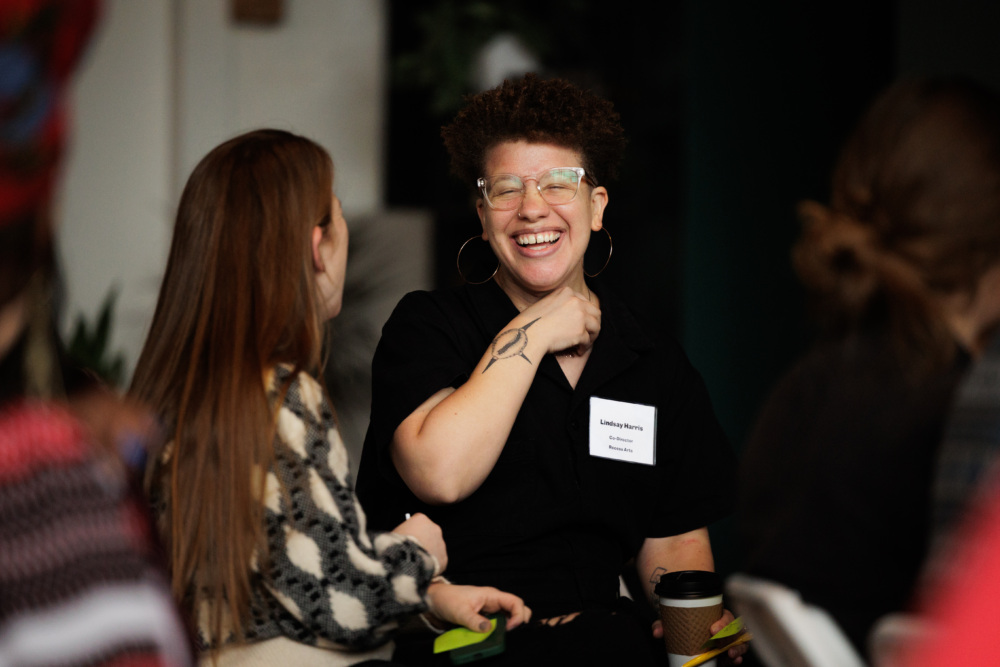 A person wearing glasses and a name tag laughs and chats with another seated person; both appear engaged in conversation at an indoor event.
