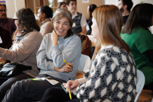 Two women sit and have a conversation during a group event, holding pens and paper, while others talk in the background.