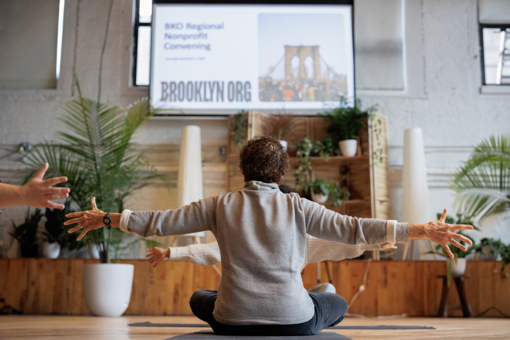 A person sits cross-legged with arms outstretched in a yoga pose indoors, with "BKD Regional Nonprofit Convening" and "BROOKLYN.ORG" displayed on a screen in the background.
