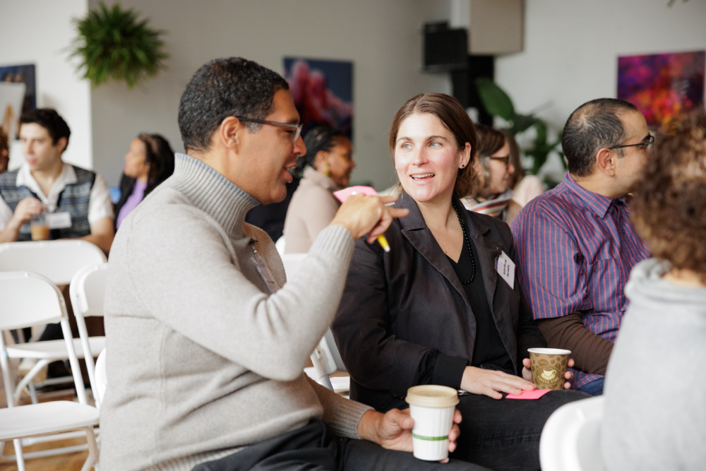 People sitting in white chairs at an indoor event, holding coffee cups and talking. Some have name tags. Plants and paintings are visible in the background.
