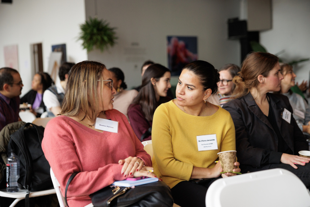 People sit in rows at an indoor event, two women in the foreground converse while holding notepads and cups, with name tags visible on their sweaters.