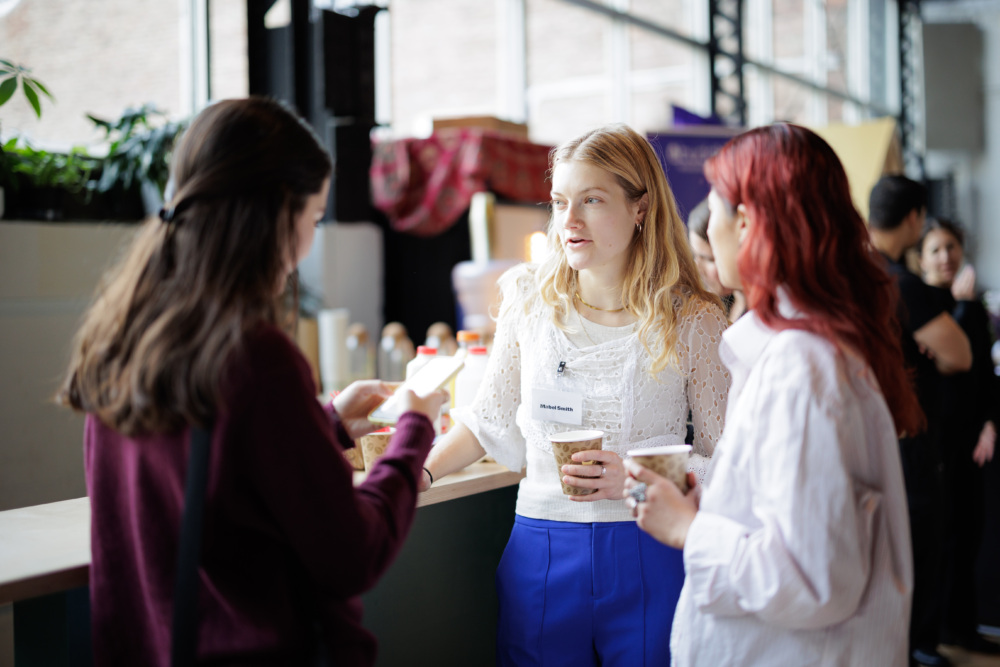 Three women stand together indoors, holding cups and conversing near a counter with food and drinks, as other people gather in the background.