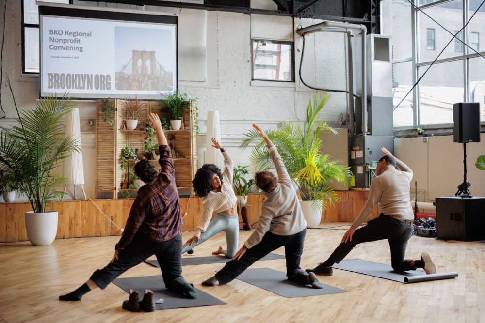 Four people on yoga mats perform a side stretch in a spacious indoor venue with plants, wooden floors, and a screen displaying “BKO Regional Nonprofit Convening.”.