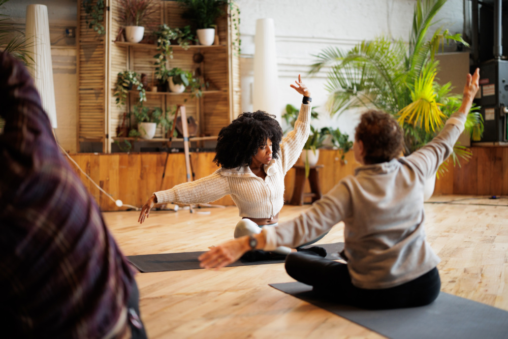 Several people sit on yoga mats in a spacious room with plants, stretching their arms out during a seated exercise class.