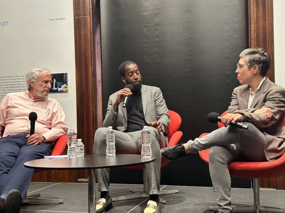 Three people sit on red chairs, holding microphones and conversing during a panel discussion. Bottled water and microphones rest on a black table in front of them.