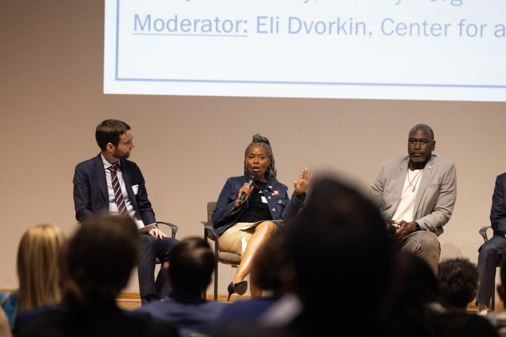Three panelists sit on stage during a moderated discussion; one woman in the center speaks into a microphone while the others listen.
