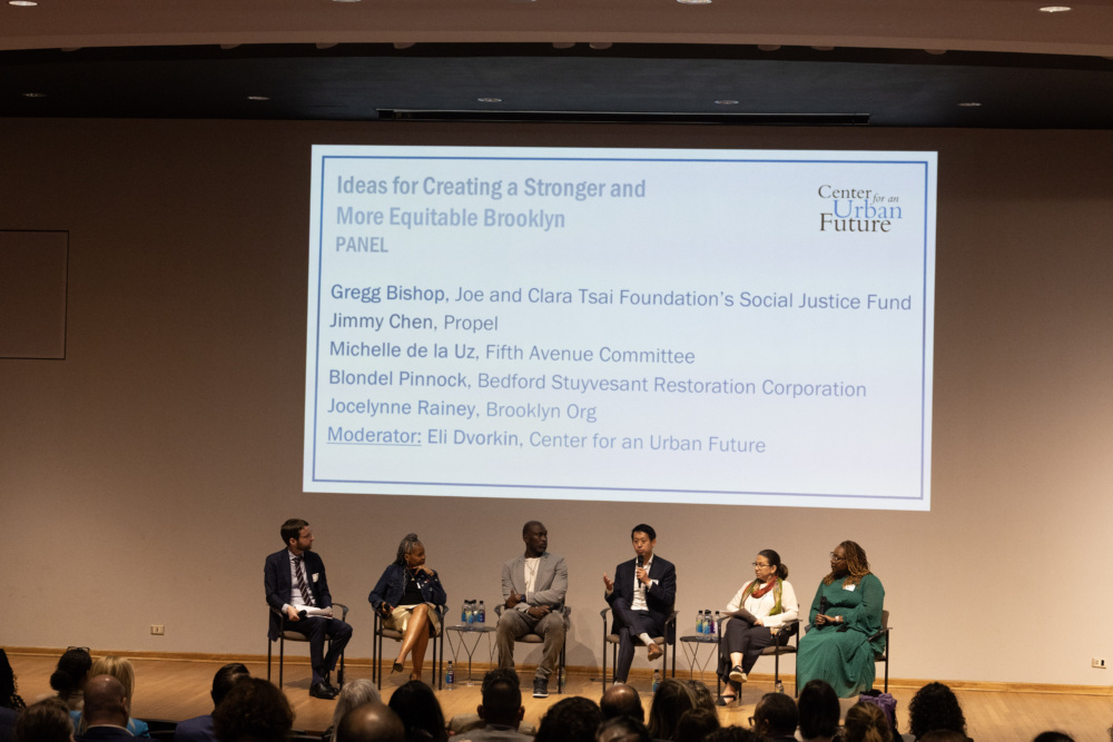 Six panelists sit onstage in front of a large screen displaying their names and affiliations at the "Ideas for Creating a Stronger and More Equitable Brooklyn" event.