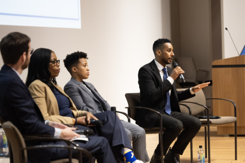 Four panelists sit on stage, one holding a microphone and speaking while the others listen, during a discussion event in a conference room.