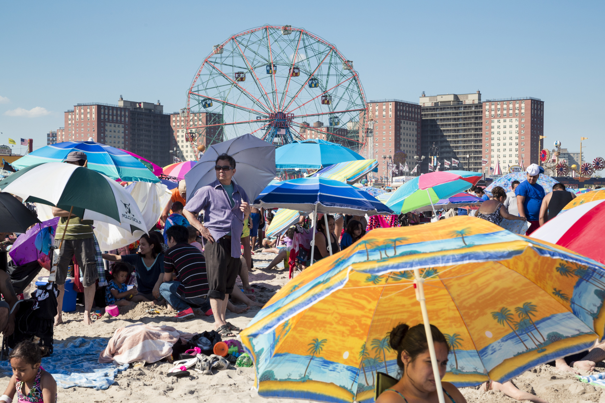 Crowded beach scene with people under colorful umbrellas, sand, and a large Ferris wheel and tall buildings visible in the background on a clear day.