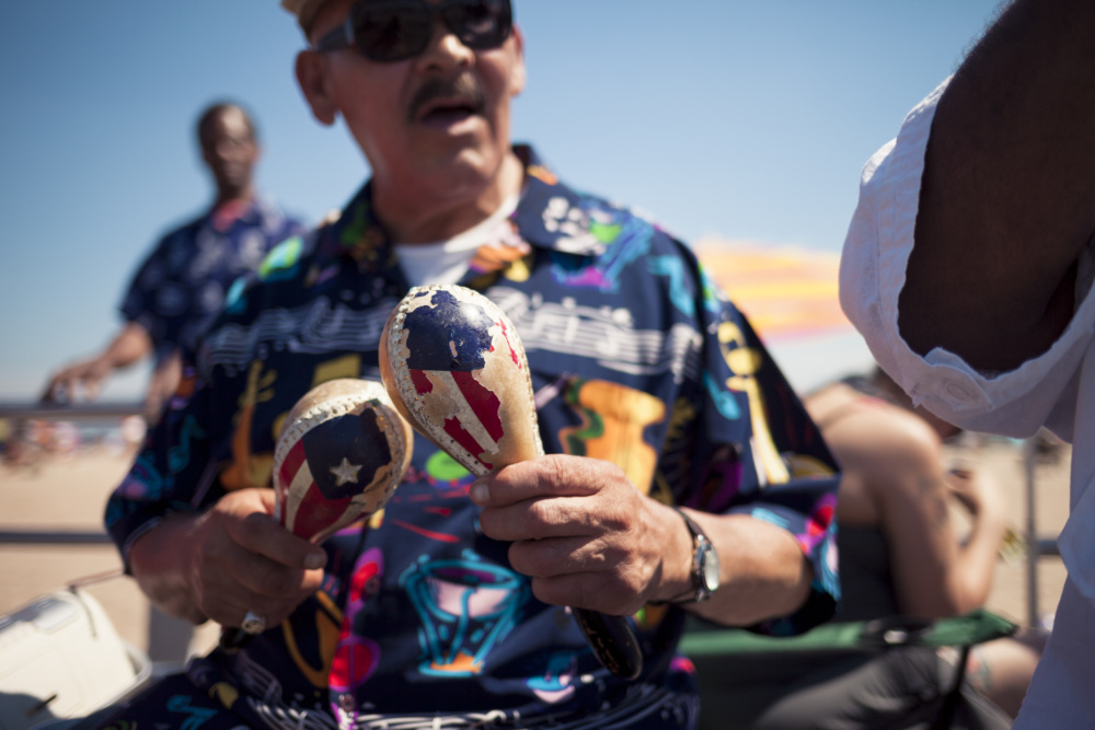 A man wearing sunglasses and a patterned shirt plays maracas painted with Puerto Rican flags outdoors on a sunny day.