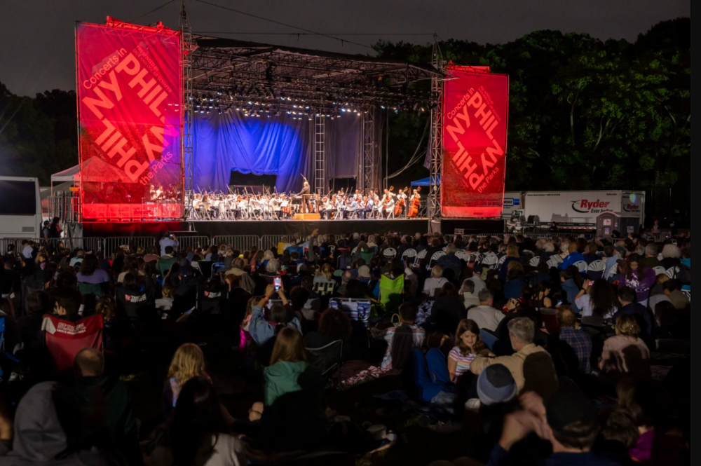 A large audience sits outdoors at night watching a live orchestra perform on a lit stage with red banners that read "PHIL." Trees and a truck are visible in the background.