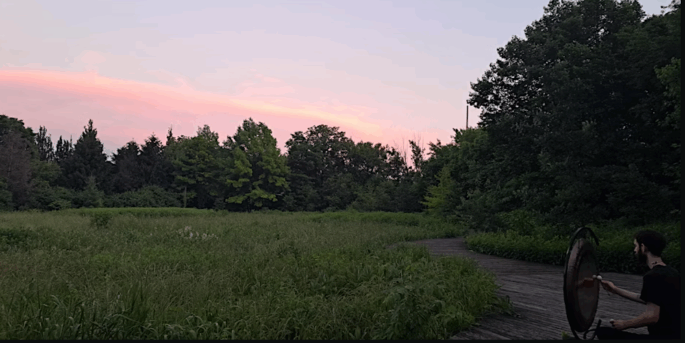 A person stands on a wooden path in a grassy field near trees at sunset, holding and striking a large gong.