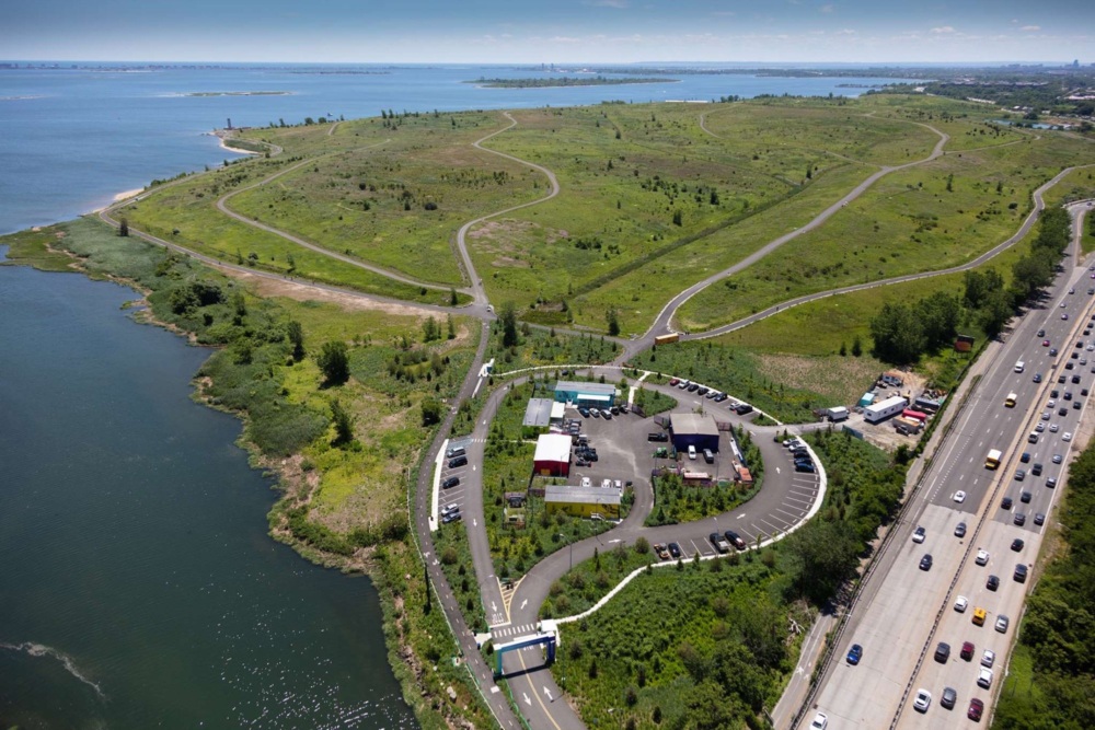 Aerial view of a green parkland area beside a body of water, with curved walking paths, a parking lot, and a busy highway on the right.