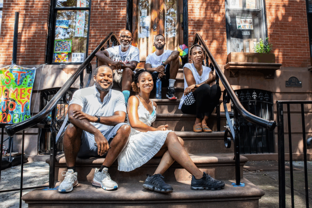 Five people sit and pose together on the steps of a brownstone building during the daytime, with sunlight and colorful welcome signs in the background.