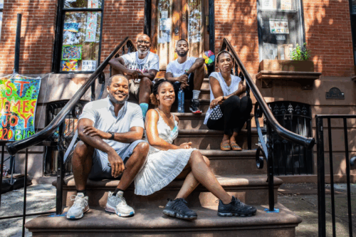 Five people sit and pose together on the steps of a brownstone building during the daytime, with sunlight and colorful welcome signs in the background.