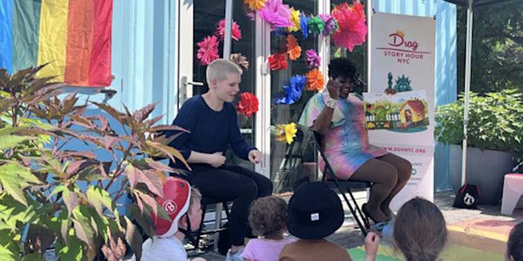 Two presenters sit in front of children outdoors, with colorful decorations, a Drag Story Hour NYC banner, and a rainbow flag hanging on the building.