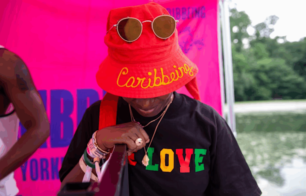 Person wearing a red bucket hat with "Caribbean" embroidered, sunglasses, and a black "LOVE" t-shirt stands outdoors in front of a pink backdrop.