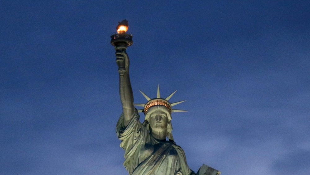 The Statue of Liberty stands illuminated against a dark, cloudy evening sky, holding a torch in her right hand and a tablet in her left.