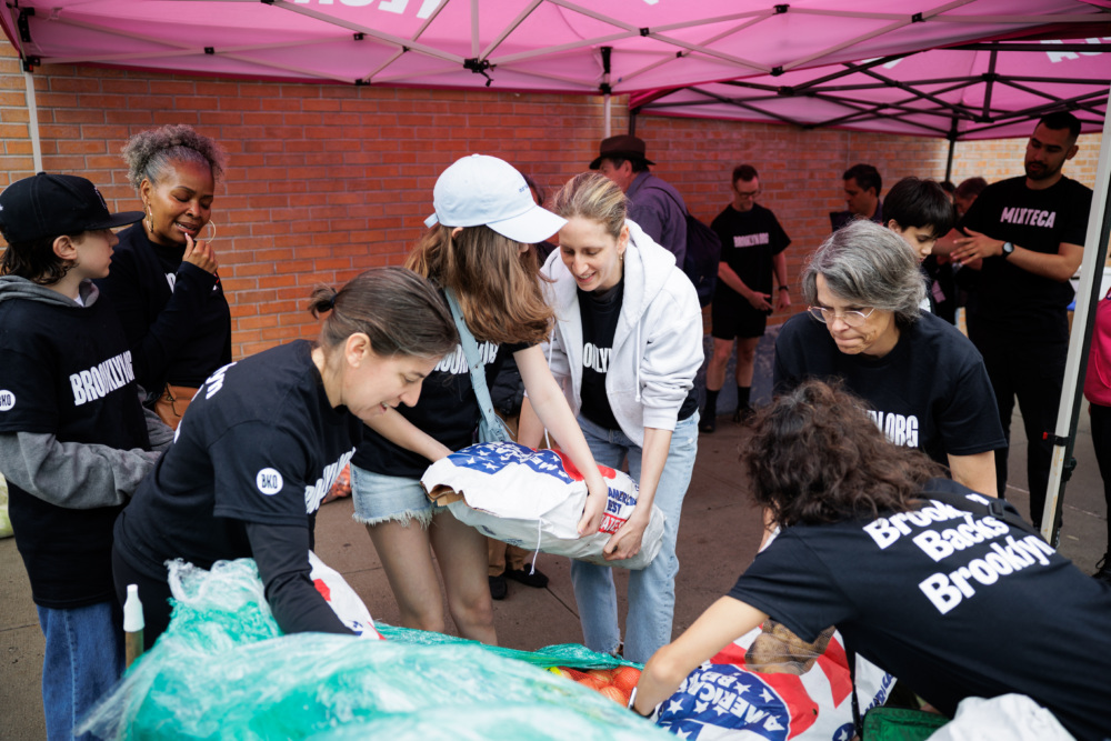 A group of volunteers wearing matching shirts work together under a canopy to sort and bag supplies, including bags with stars and stripes, near a brick wall.