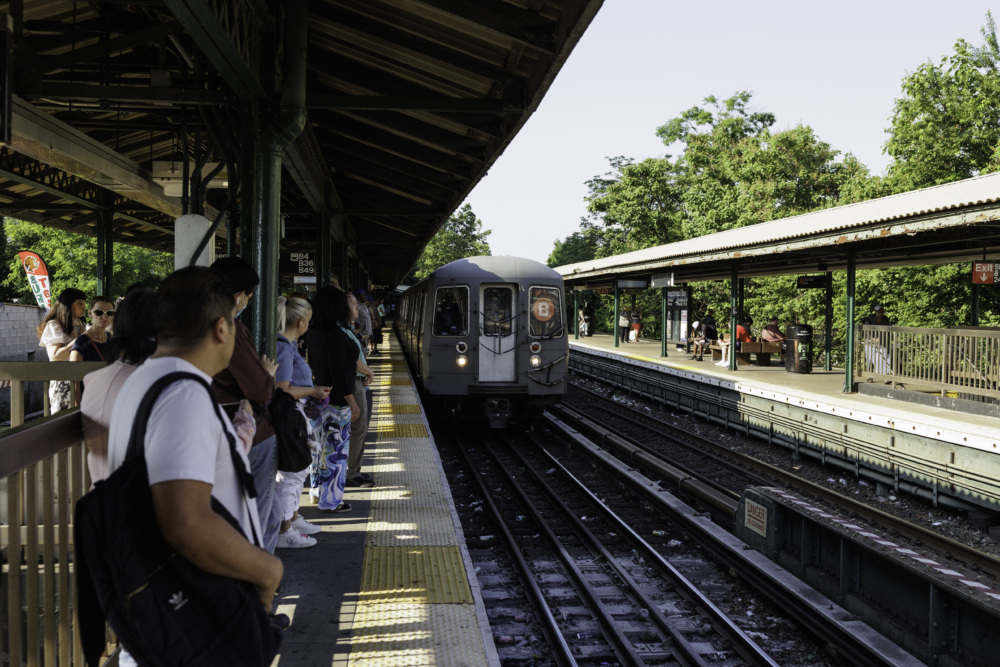 A subway train approaches an outdoor station platform as a group of people wait along the edge on a sunny day.