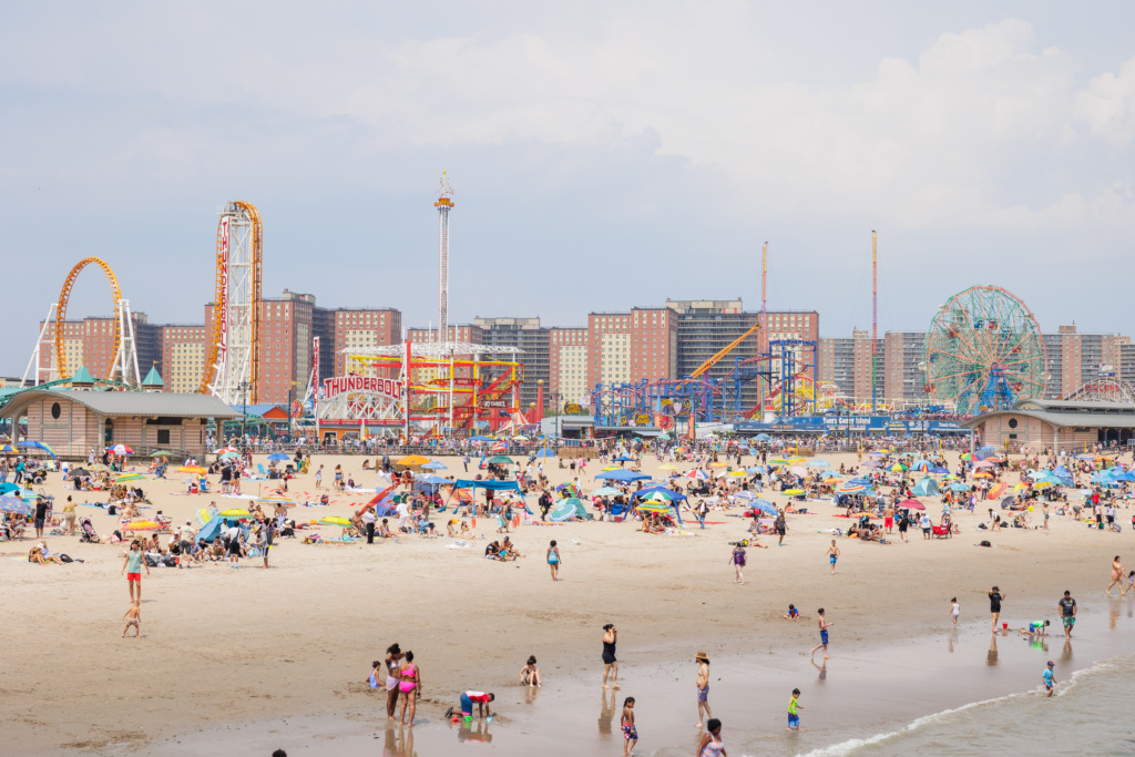Crowded beach with people swimming, sunbathing, and playing in the sand; amusement park rides and city buildings are visible in the background.