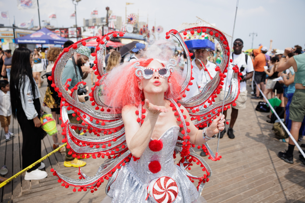 A person in a silver costume with red, white, and pom-pom decorations blows a handful of powder on a crowded boardwalk during a festive event.